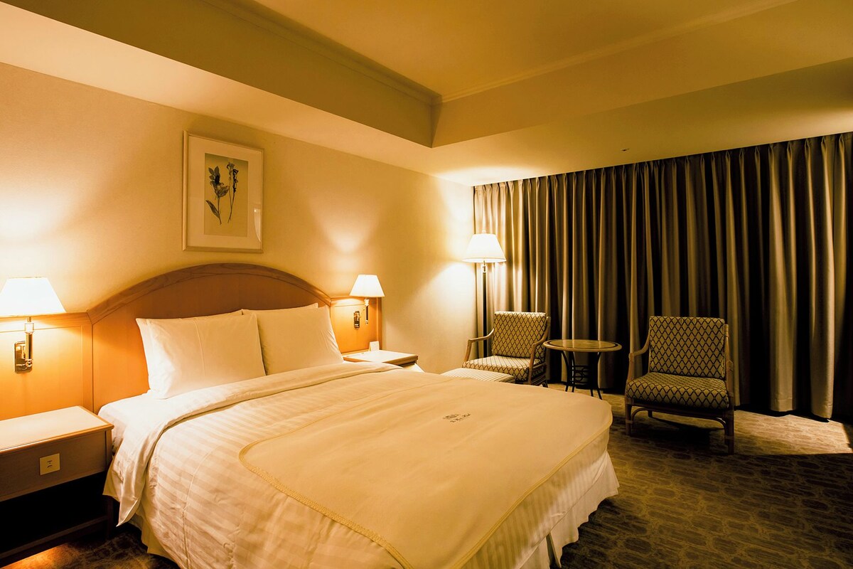 Een hotelkamer met een bed, twee bedlampen, een nachtkastje, een stoel, een klein tafeltje en een groot raam met gordijnen.