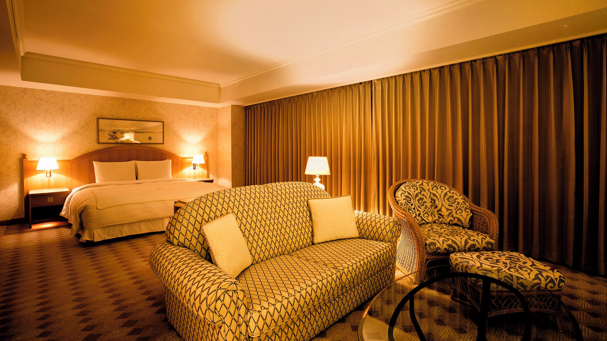 A hotel room with a bed, two bedside lamps, a patterned sofa, a patterned armchair, and a large window with curtains.