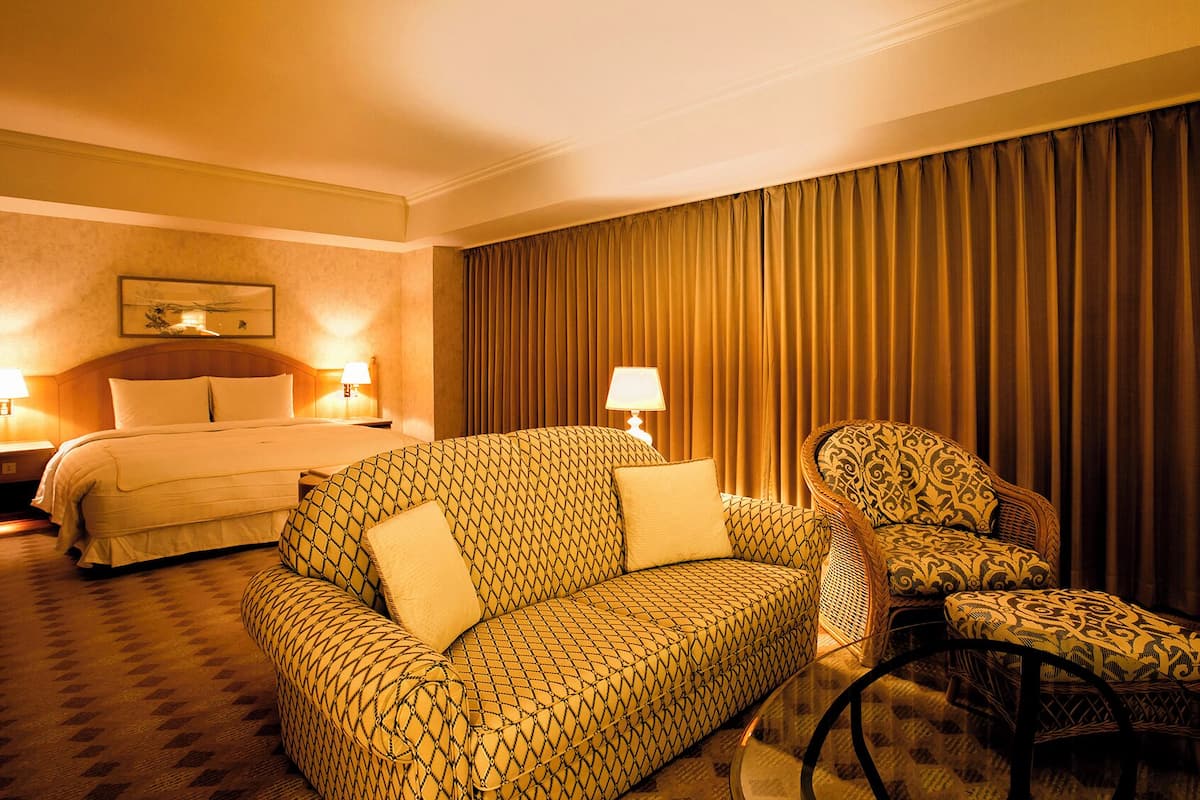 Une chambre d’hôtel avec un lit, deux lampes de chevet, un canapé à motifs, un fauteuil à motifs et une grande fenêtre avec des rideaux.