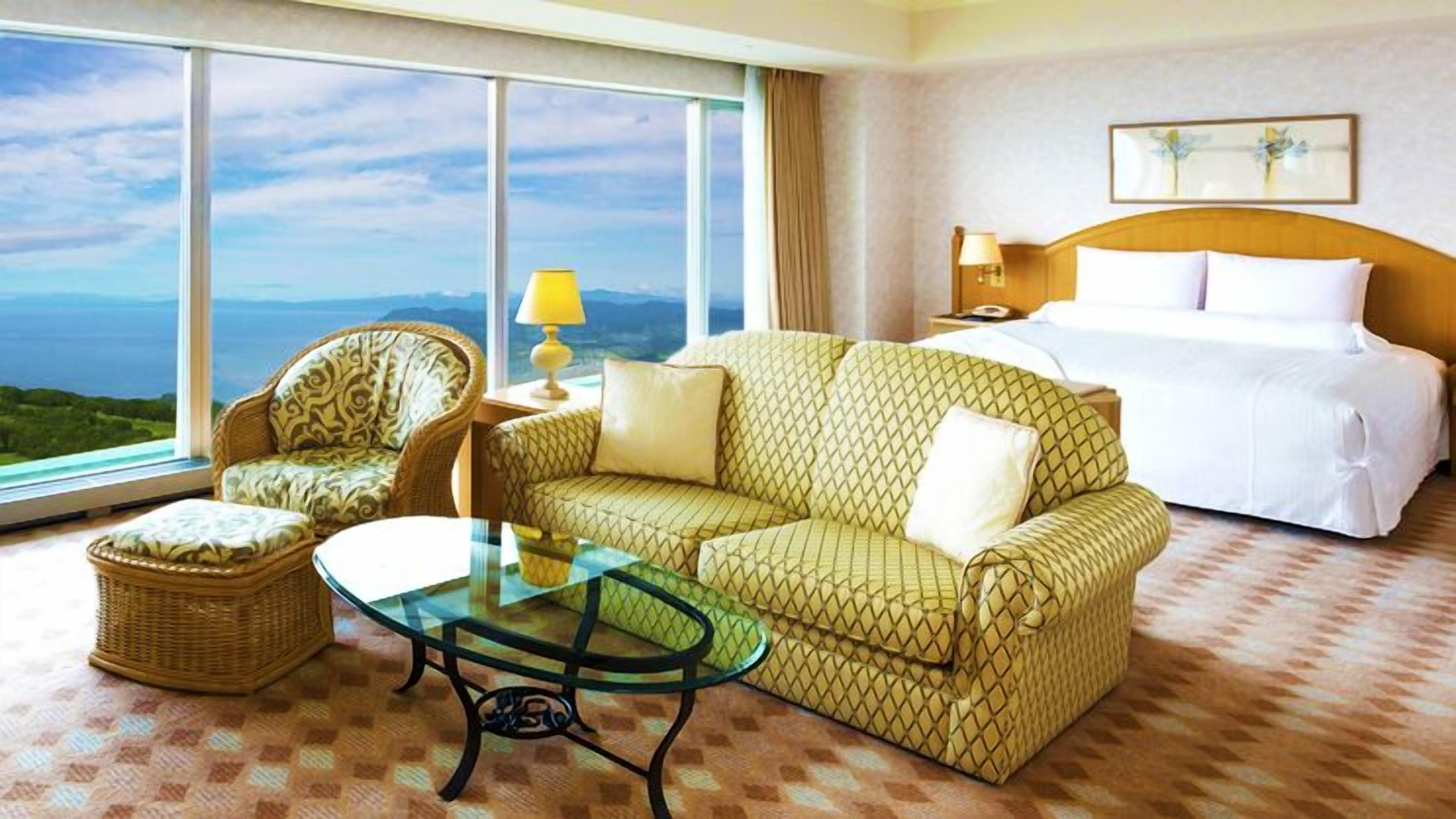 Ein Hotelzimmer mit Bett, Sofa, Sessel, einem Glastisch und Blick auf die Landschaft.