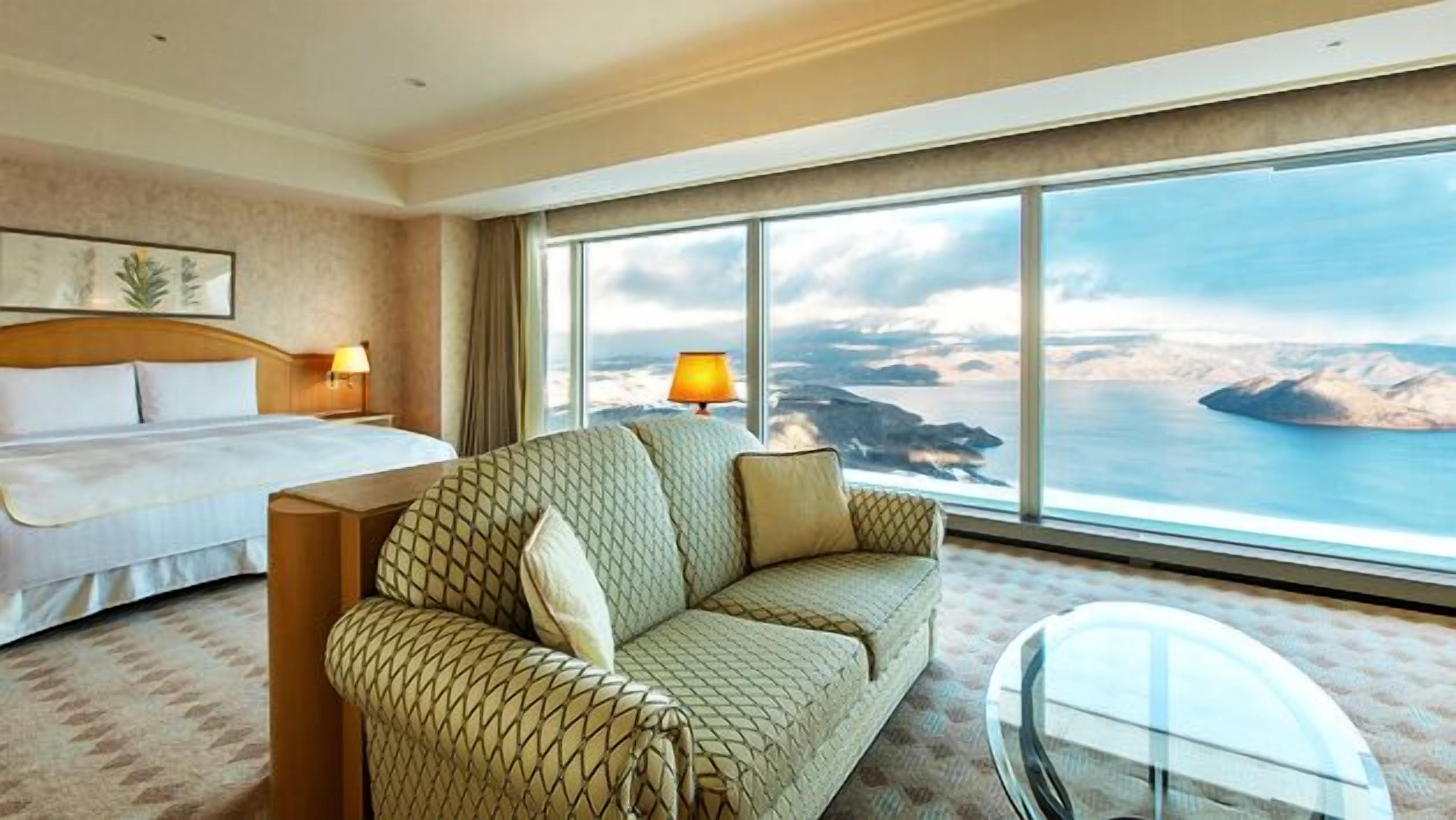 Ein Hotelzimmer mit Bett, einer Couch, einem Glastisch und einem großen Fenster mit Blick auf eine Küstenlandschaft.