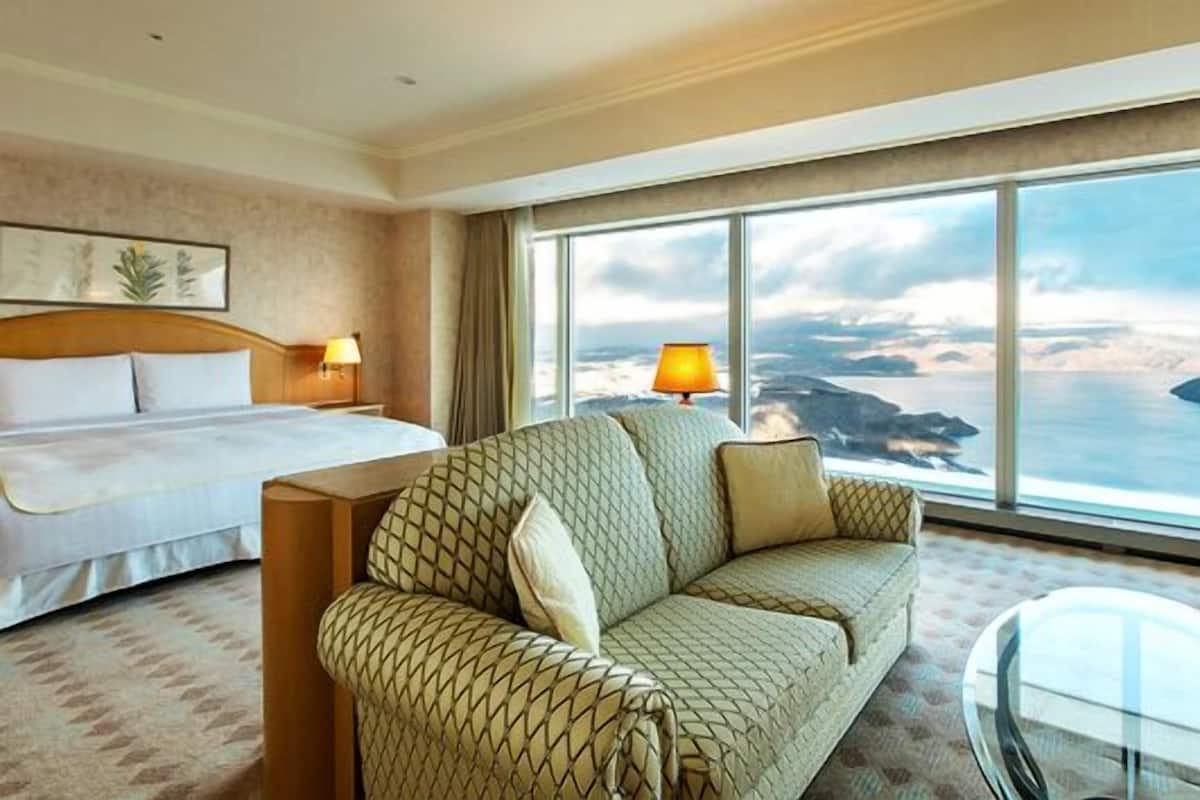 Une chambre d’hôtel avec un lit, un canapé, une table en verre et une grande fenêtre donnant sur un paysage côtier.