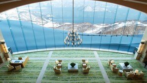 Een kamer met een groot rondboogvenster, een kroonluchter en meerdere zithoeken met tafels en stoelen.