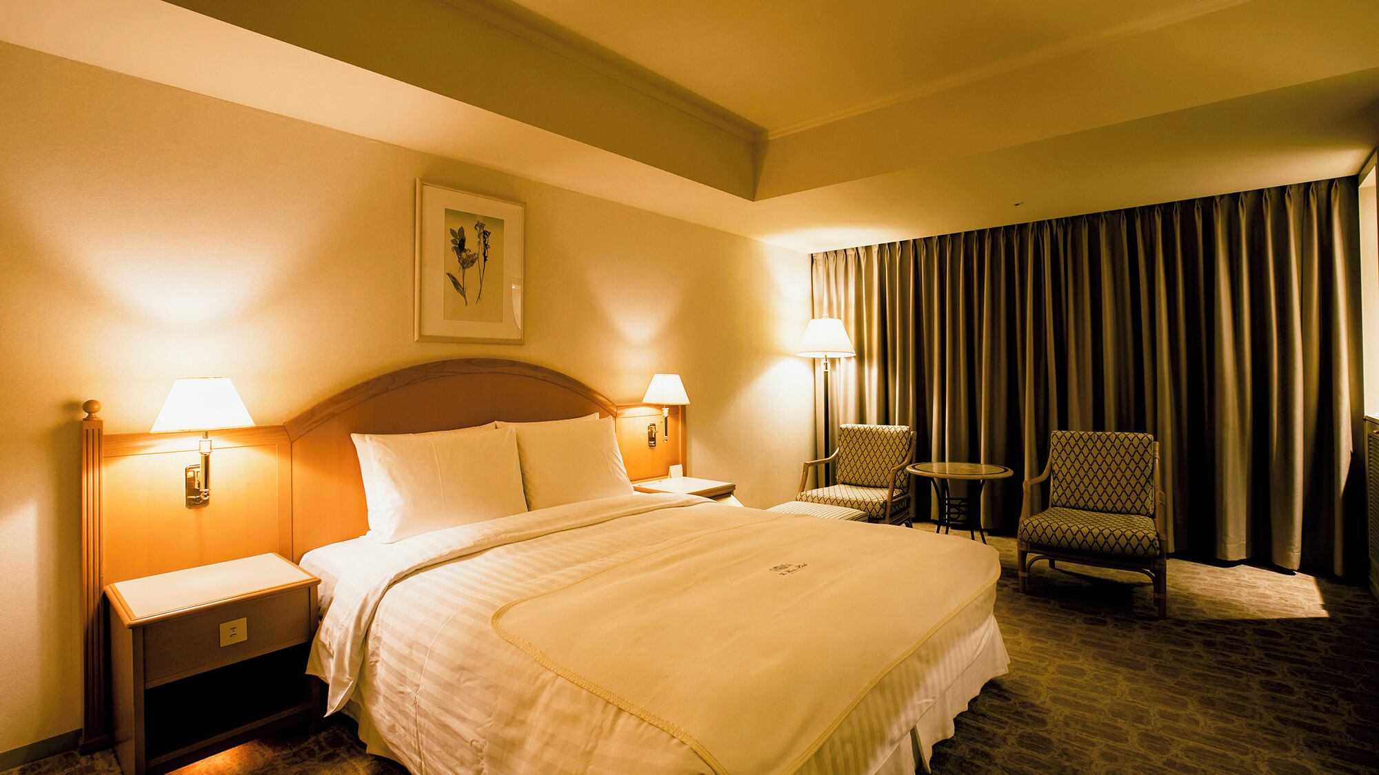 A hotel room with a bed, two bedside lamps, a nightstand, a chair, a small table, and a large window with curtains.