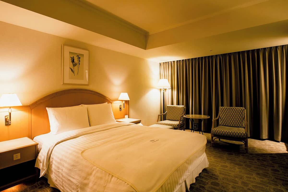Une chambre d’hôtel comprenant un lit, deux lampes de chevet, une table de chevet, une chaise, une petite table et une grande fenêtre avec des rideaux.