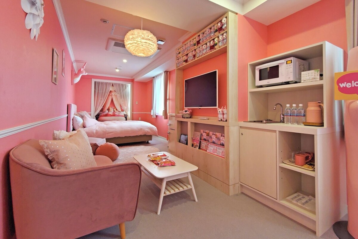 A pink-themed hotel room with a bed, a sofa, a small table, a TV, a microwave, and a 'welcome!' sign.