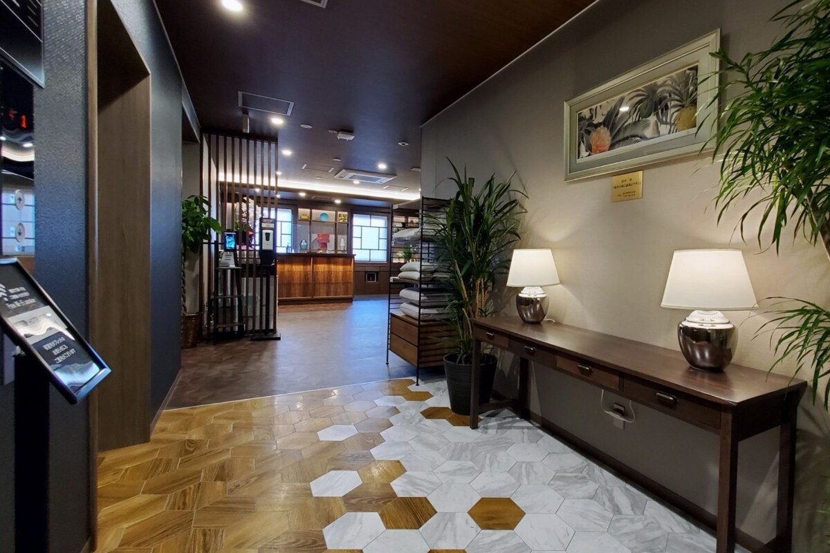 A hotel lobby with a reception desk, a wooden floor with hexagonal tiles, a reception area with a counter and chairs, and a large potted plant.