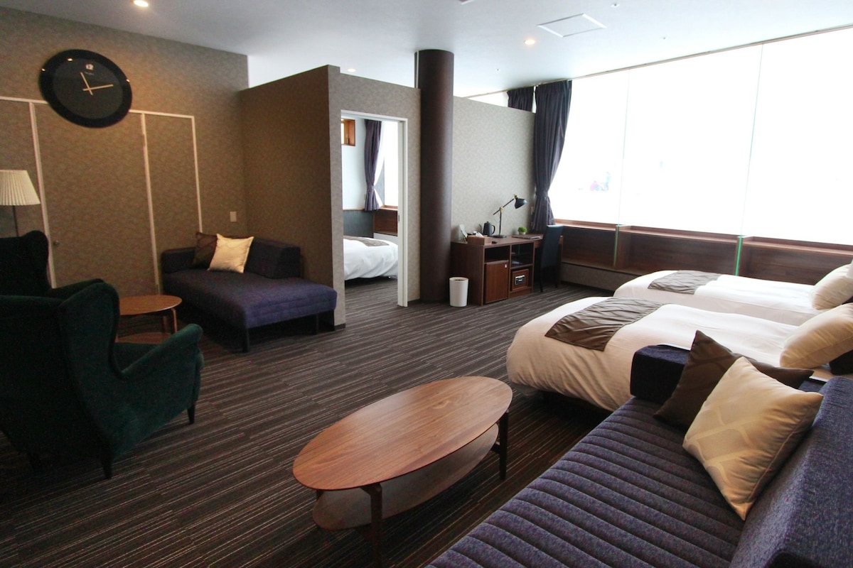 A hotel room with a sofa, two beds, a desk, a chair, a clock, and a window with curtains.