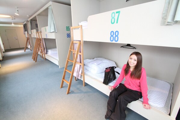 Une chambre de dortoir avec des lits superposés numérotés 87 et 88, une échelle en bois et un sac noir posé sur l’un des lits.