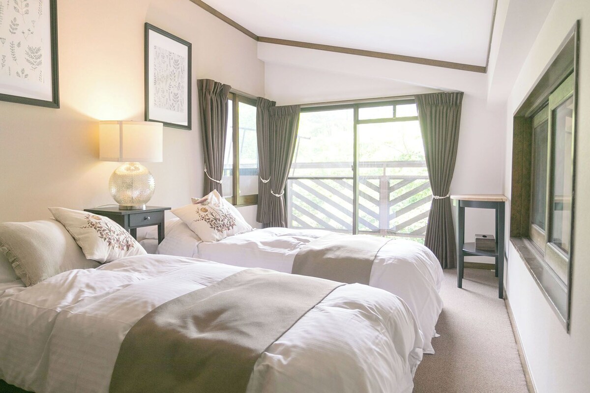 A hotel room with two beds, a nightstand with a lamp, a framed botanical print, and a window with curtains.