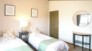 Hotellihuone, jossa on kaksi sänkyä, yöpöytä lampun kanssa, pieni pyöreä pöytä, peili ja kaksi kehystettyä kasvitaiuluista seinällä.