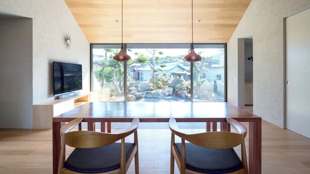 Un espace repas moderne avec une table et des chaises en bois, un téléviseur à écran plat posé sur un meuble, et une vue sur un jardin grâce à de grandes fenêtres.