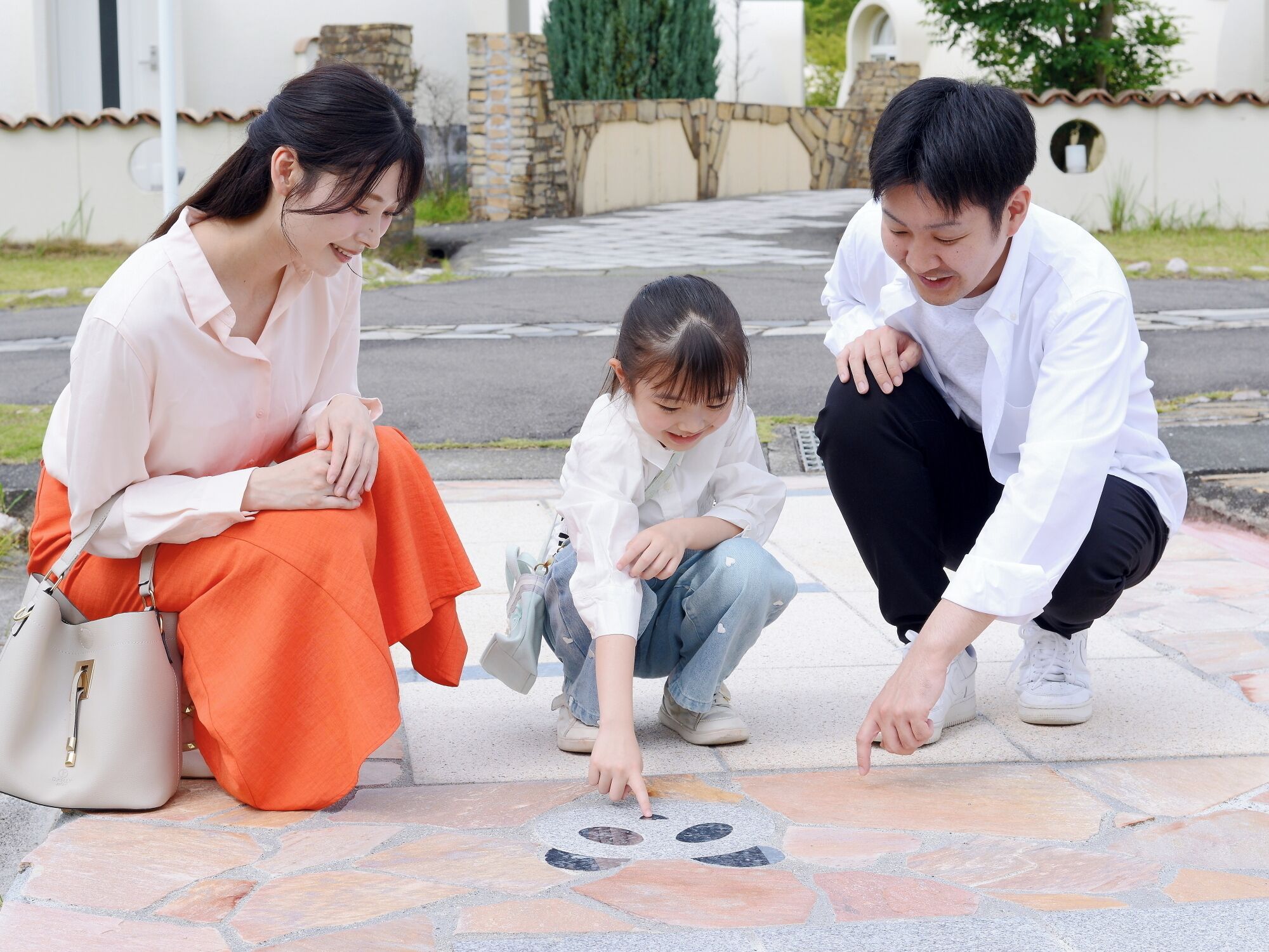 Three people sitting on a tiled outdoor area with a stone wall and greenery in the background.