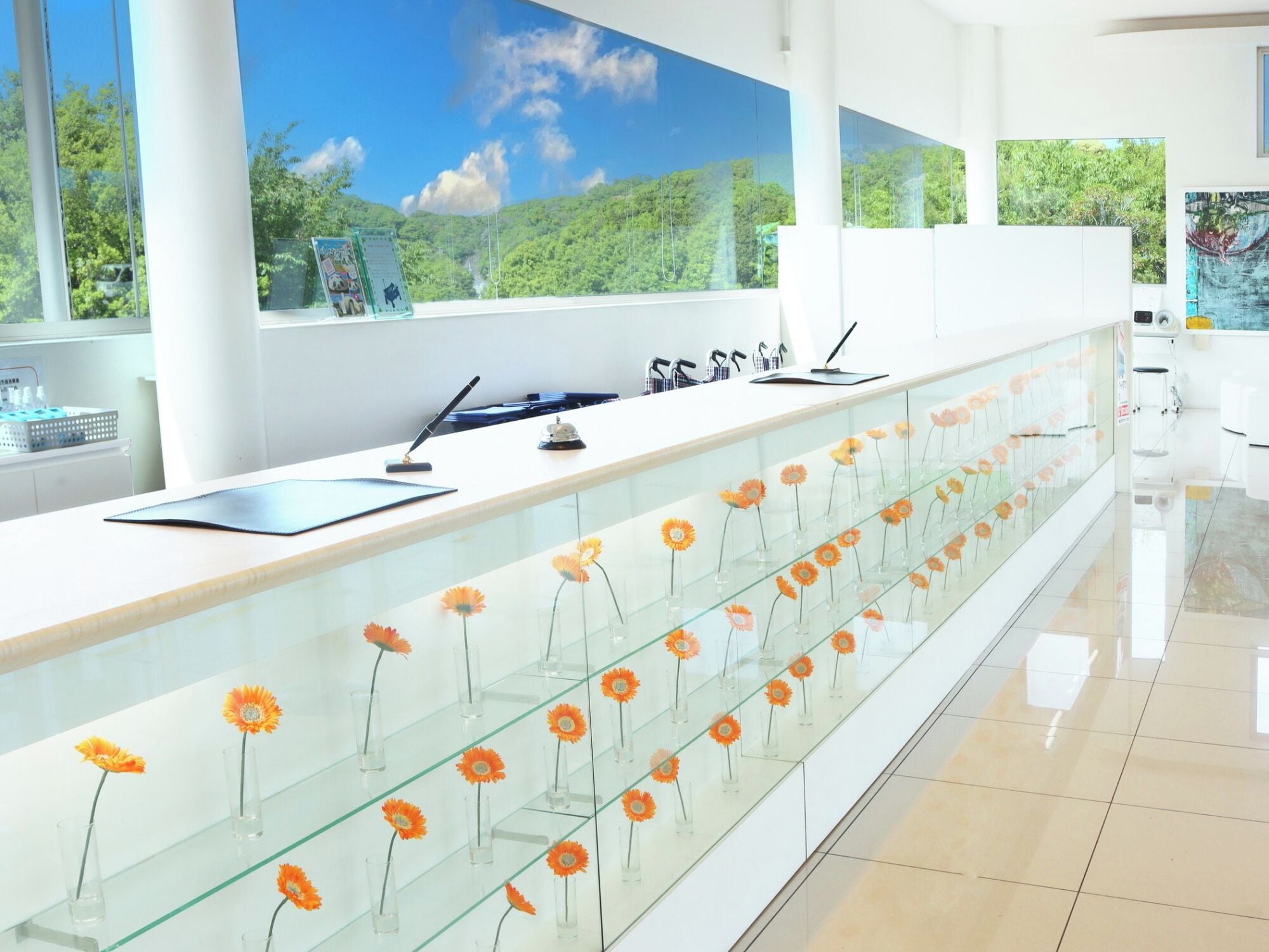 Een moderne receptiebalie met een glazen vitrine vol oranje bloemen, een witte toonbank en uitzicht op groen door grote ramen.
