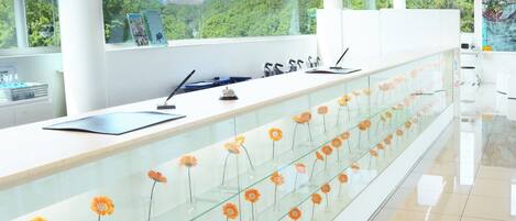 Een moderne receptiebalie met een glazen vitrine vol oranje bloemen, een witte toonbank en uitzicht op groen door grote ramen.