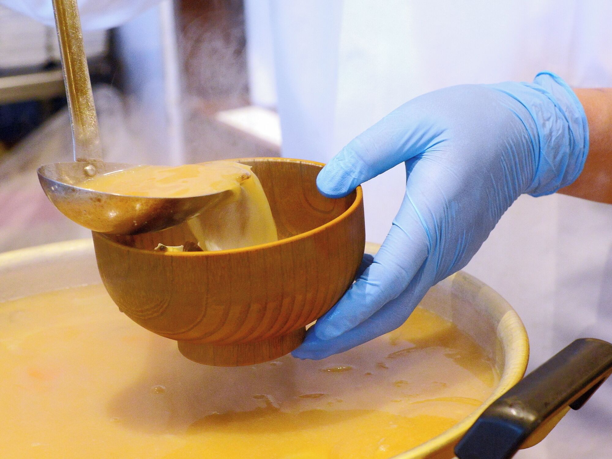 A person wearing a blue glove holding a wooden bowl over a pan with a yellow liquid.