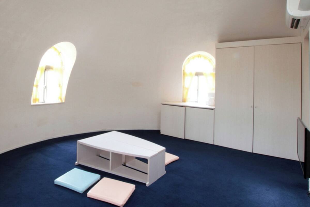 A room with a curved ceiling, white walls, a white cabinet, a small table with drawers, and two windows with arched tops.
