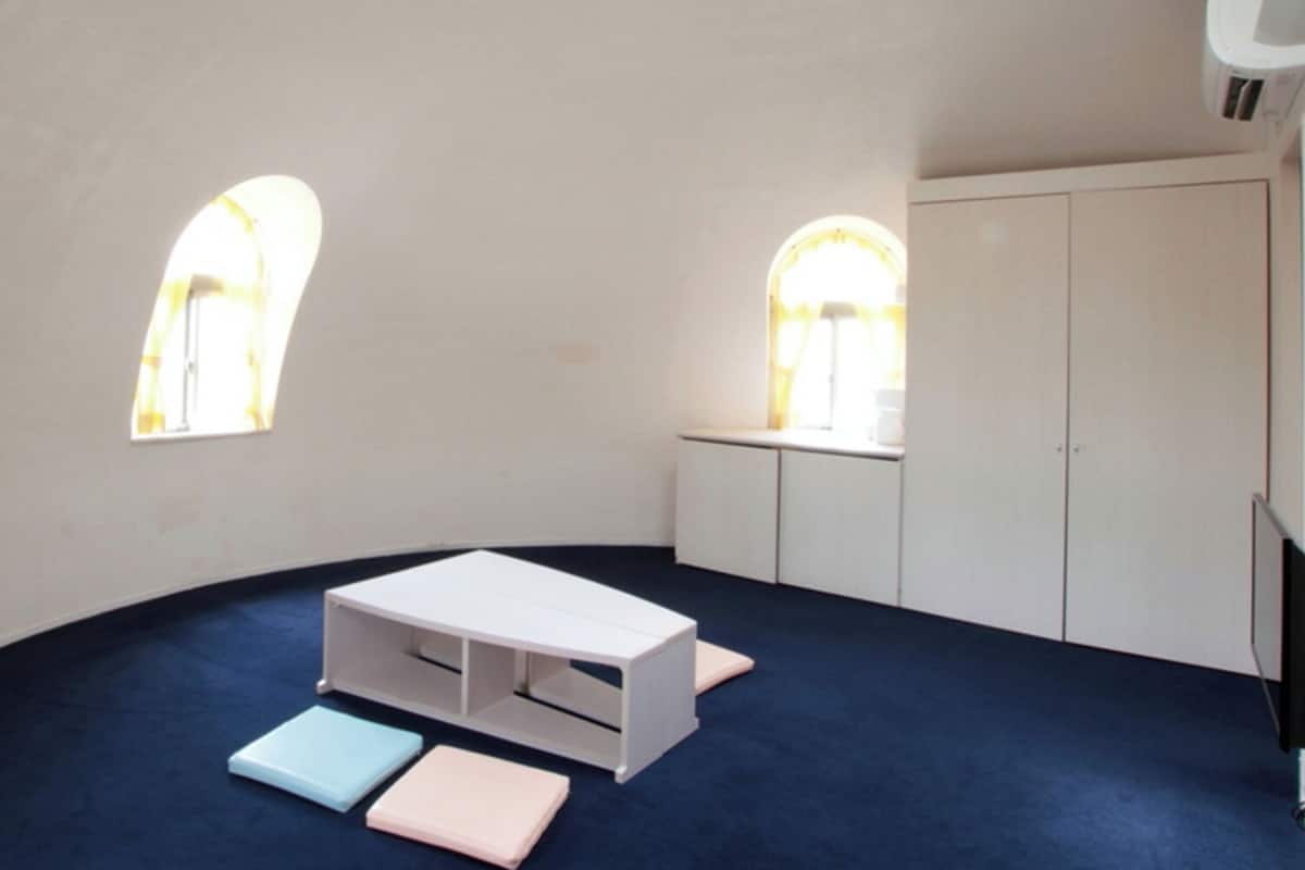 Ein Zimmer mit gewölbter Decke, weißen Wänden, einem weißen Schrank, einem kleinen Tisch mit Schubladen und zwei Fenstern mit Rundbögen.