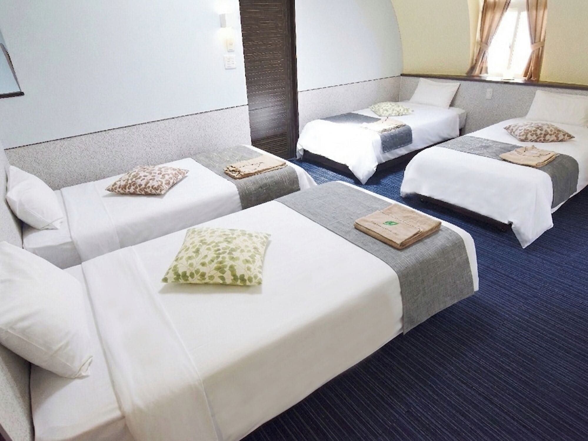 A hotel room with three beds arranged in a row, each with a pillow and a folded towel.