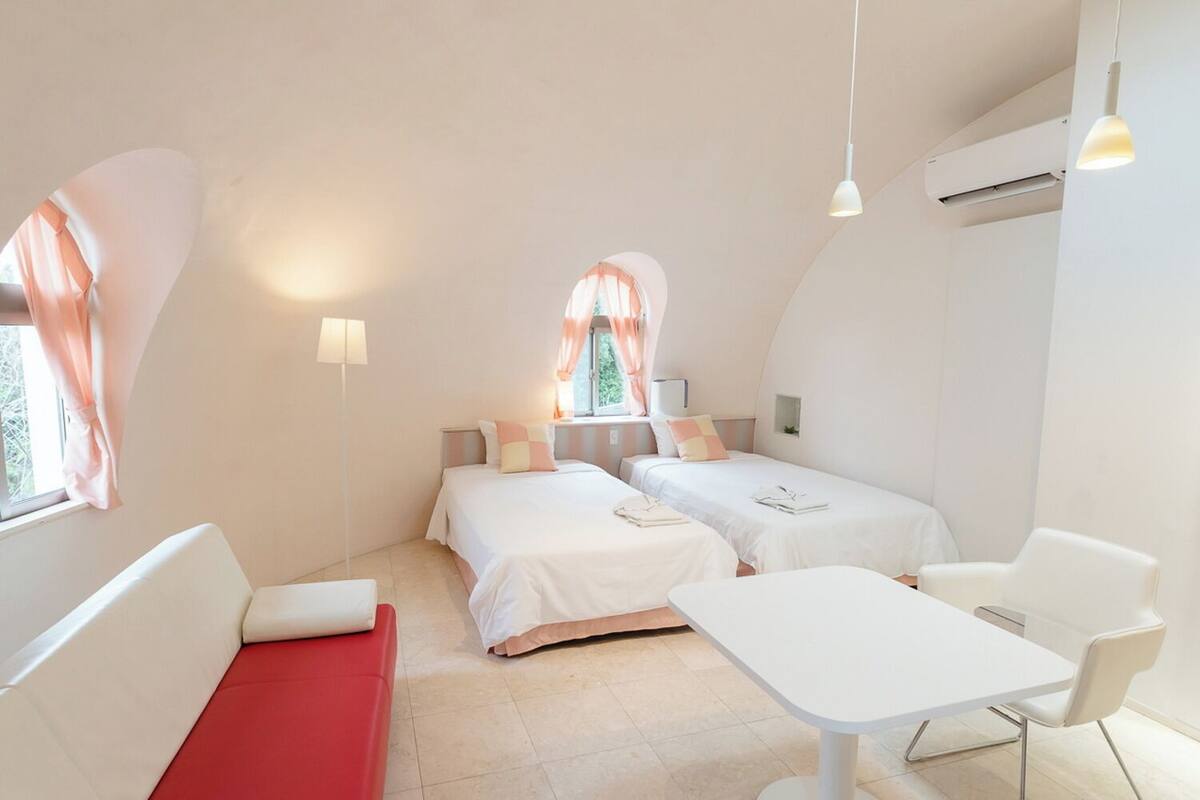 A bedroom with two beds, a red couch, a white table, and a window with curtains.