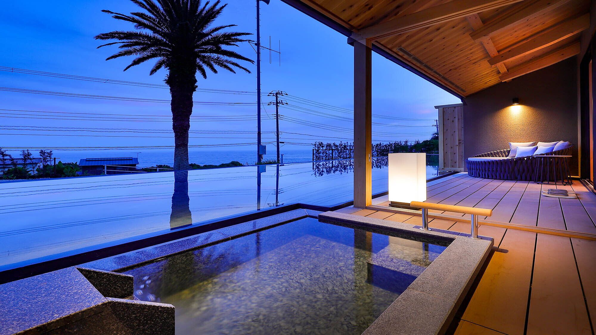 Un espace piscine extérieur moderne, doté d’une terrasse en bois, d’un coin salon avec des coussins et d’une vue sur l’océan.