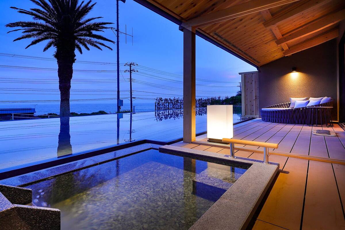Un espace piscine extérieur moderne, doté d’une terrasse en bois, d’un coin salon avec des coussins et d’une vue sur l’océan.