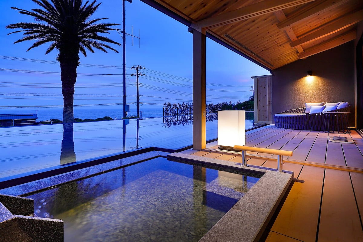 A modern outdoor pool area with a wooden deck, a seating area with cushions, and a view of the ocean.