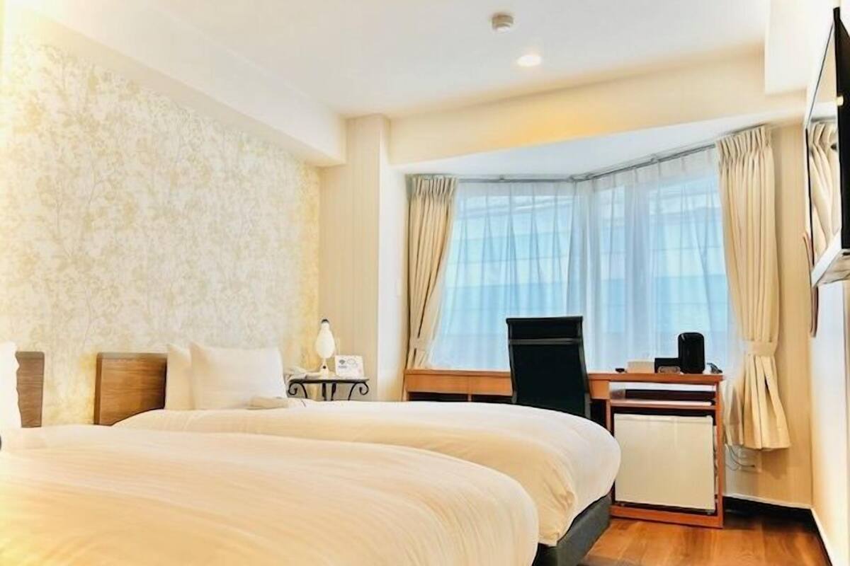 A hotel room with two beds, a desk, a chair, and a large window with curtains.