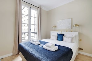 Une chambre d’hôtel avec un lit, une fenêtre avec des rideaux, un tableau au mur et une lampe.