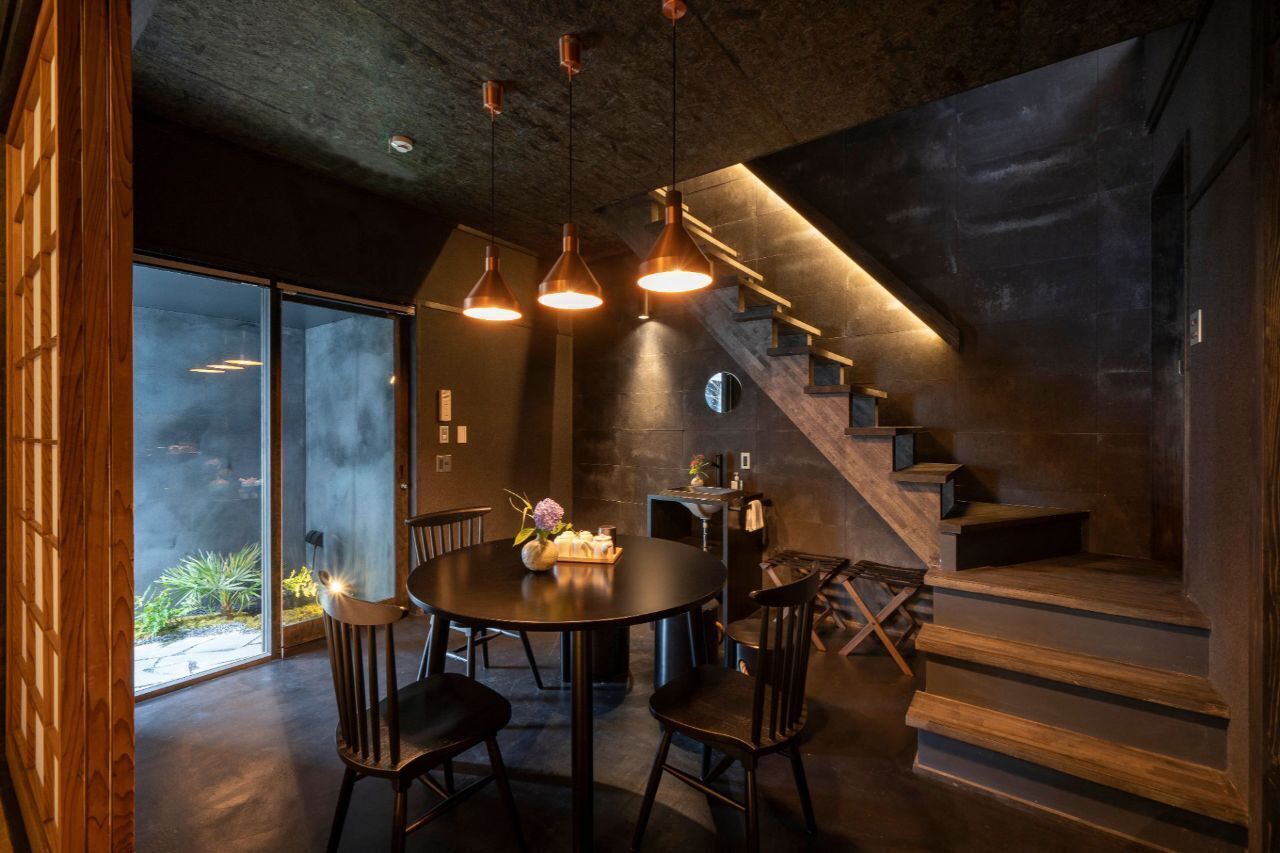 A modern interior with a round table, chairs, pendant lights, a staircase, and a view through large glass doors.