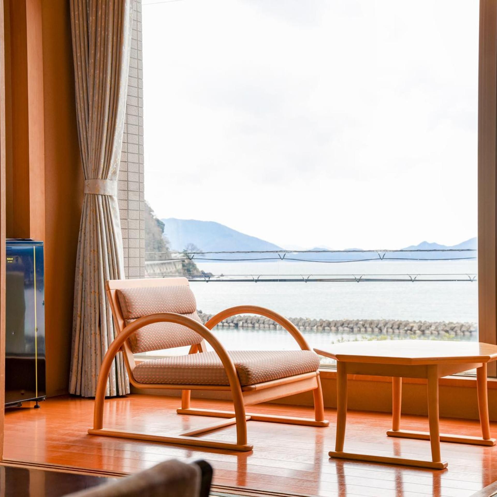 A room with a view of a body of water and mountains, featuring a wicker chair, a wooden table, and a curtain.