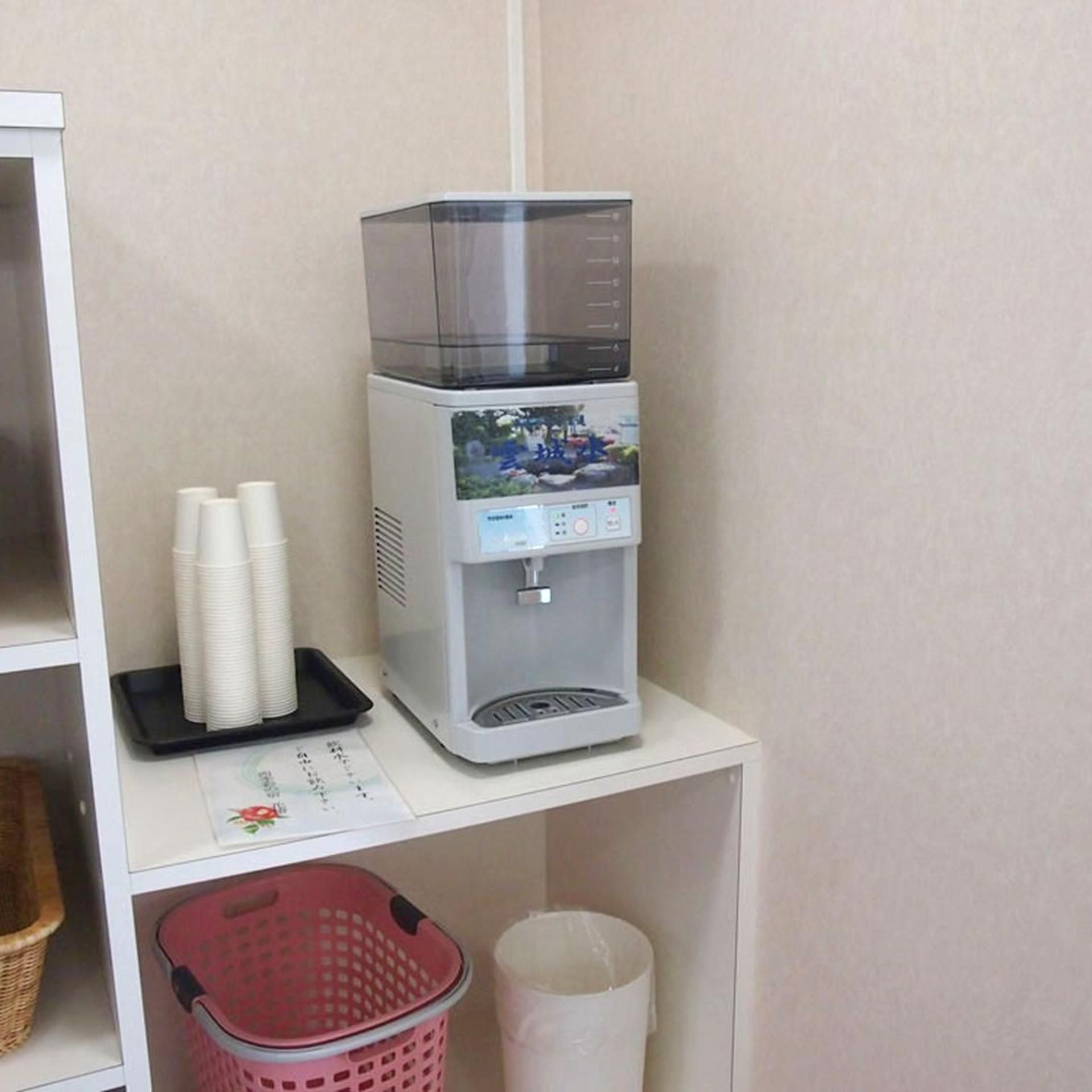 A water dispenser on a white shelf with paper cups and a red basket below.