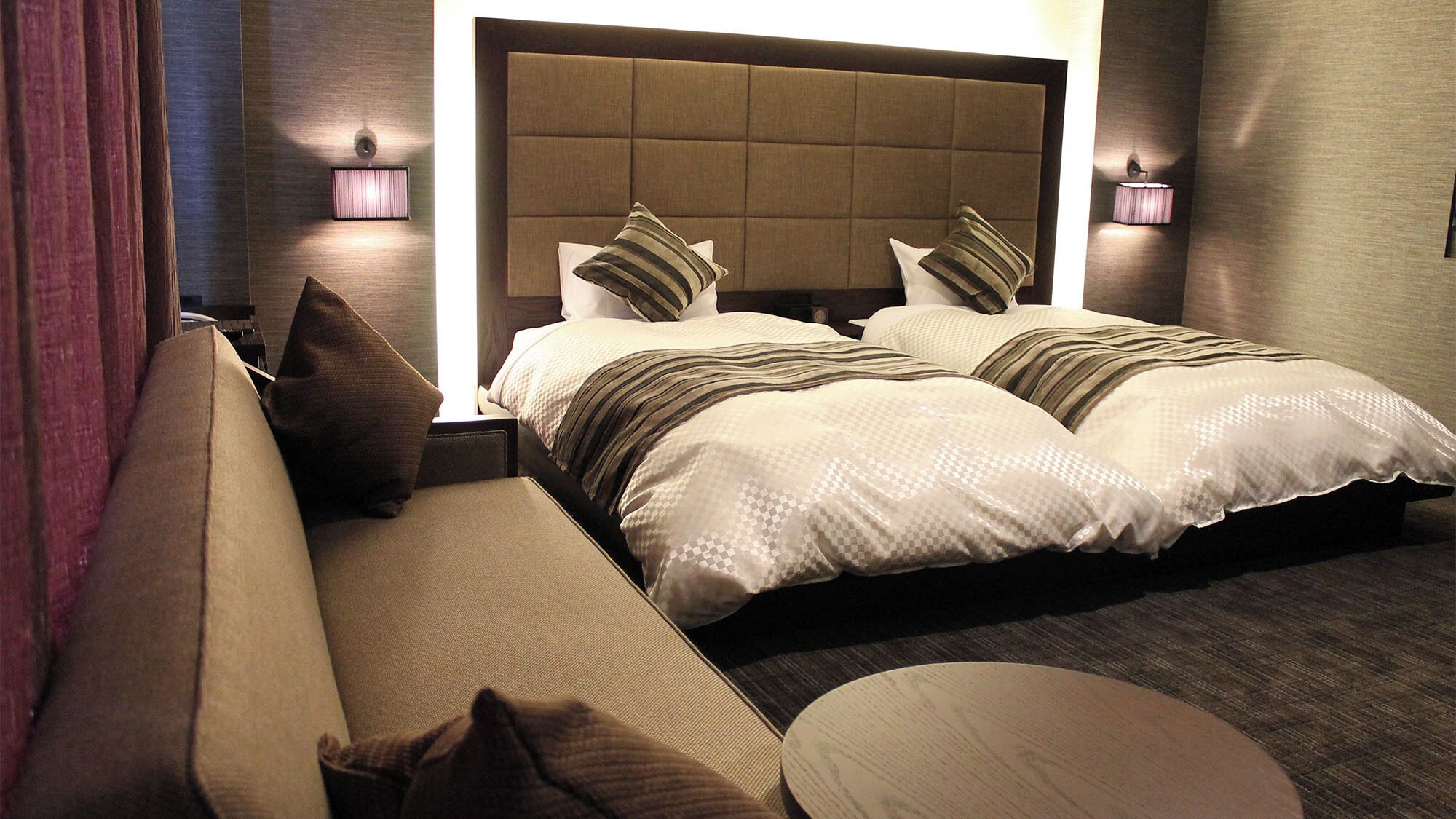 A hotel room with a large bed, a sofa, a round table, and two wall-mounted lamps.