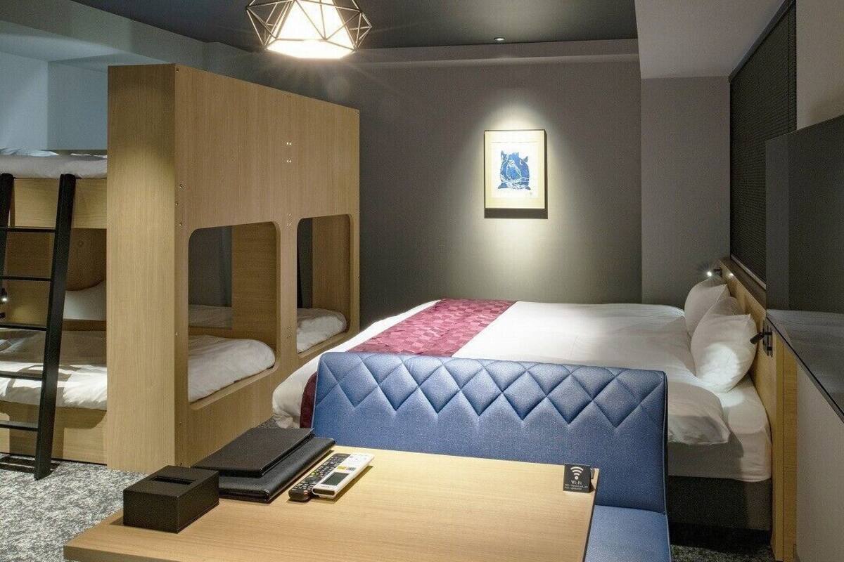 A hotel room with bunk beds, a desk, a chair, a bed with a blue headboard, a flat-screen TV, and a framed picture on the wall.