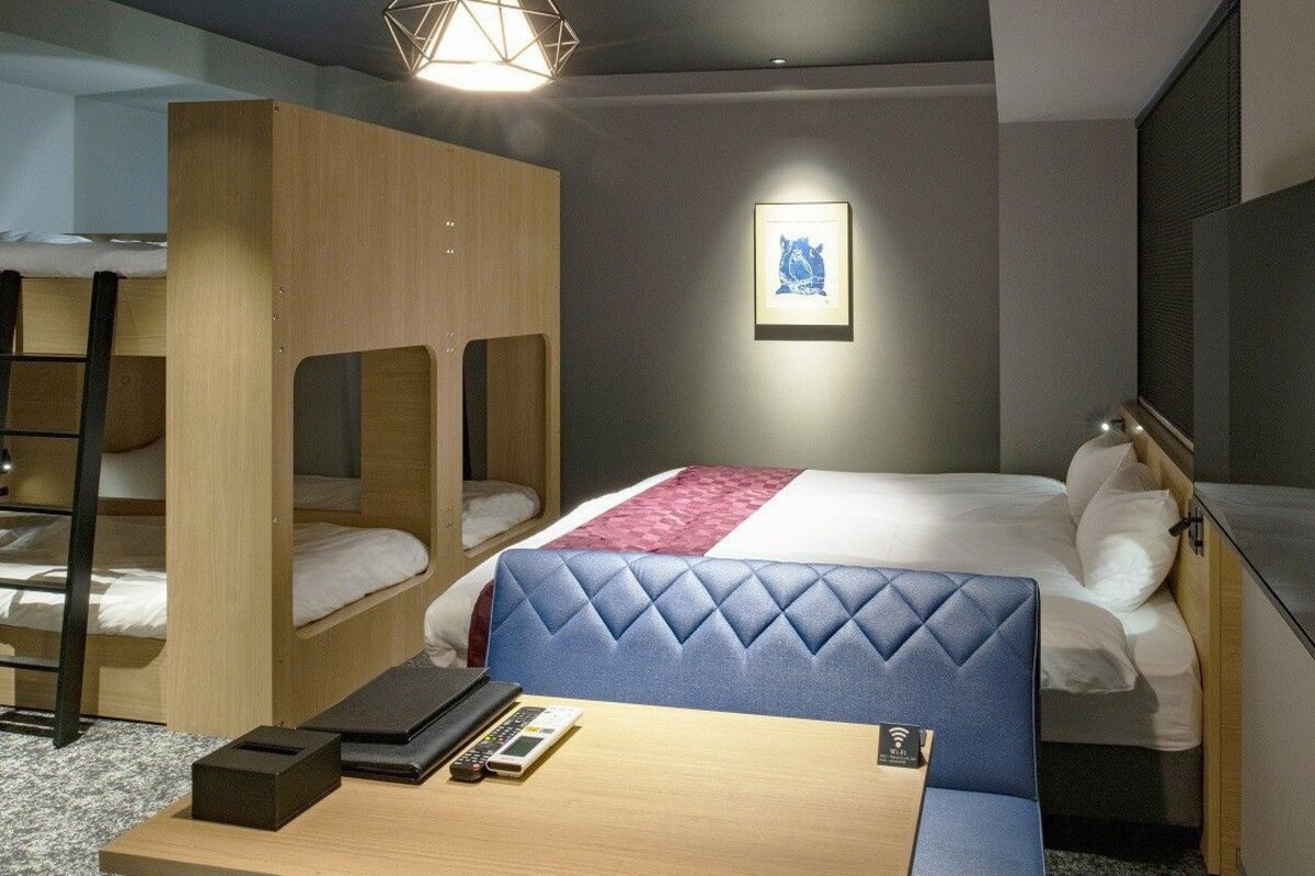 A hotel room with bunk beds, a desk, a chair, a bed with a blue headboard, a flat-screen TV, and a framed picture on the wall.