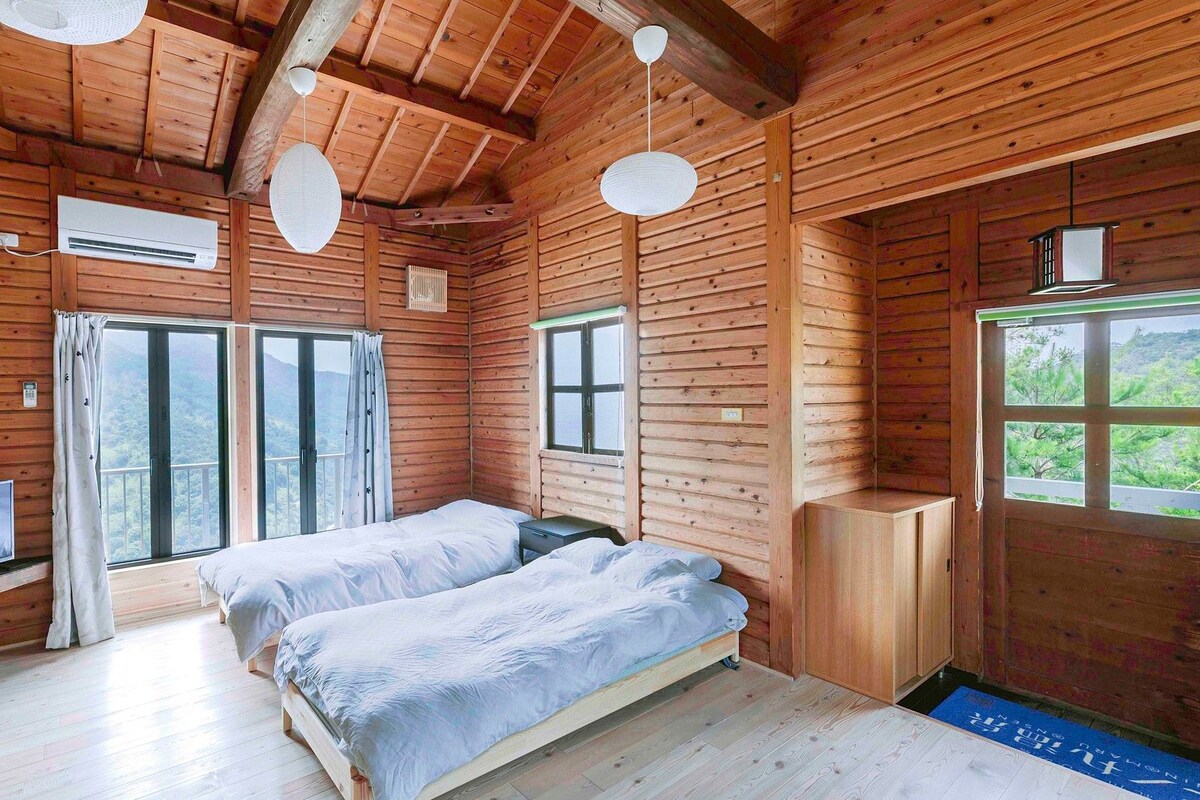 A wooden cabin room with two beds, a TV, and large windows offering a view of the outdoors.