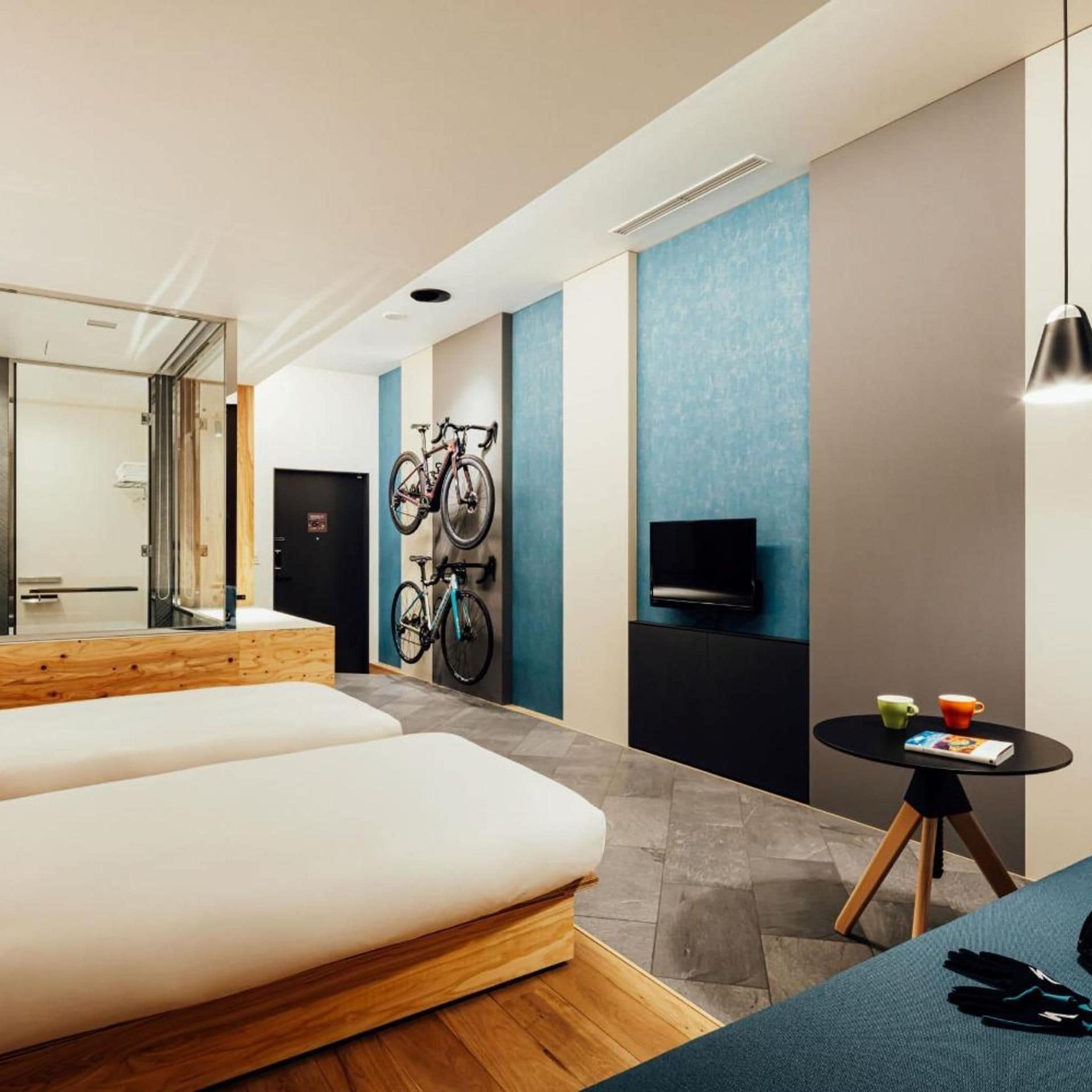 A modern hotel room with a large bed, a TV, a wall-mounted bike rack, and a small table with cups and a book.
