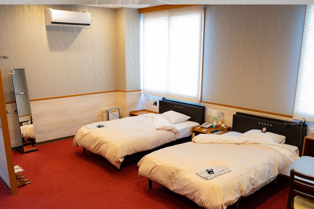 A hotel room with two beds, a desk, a chair, a mirror, and a window.