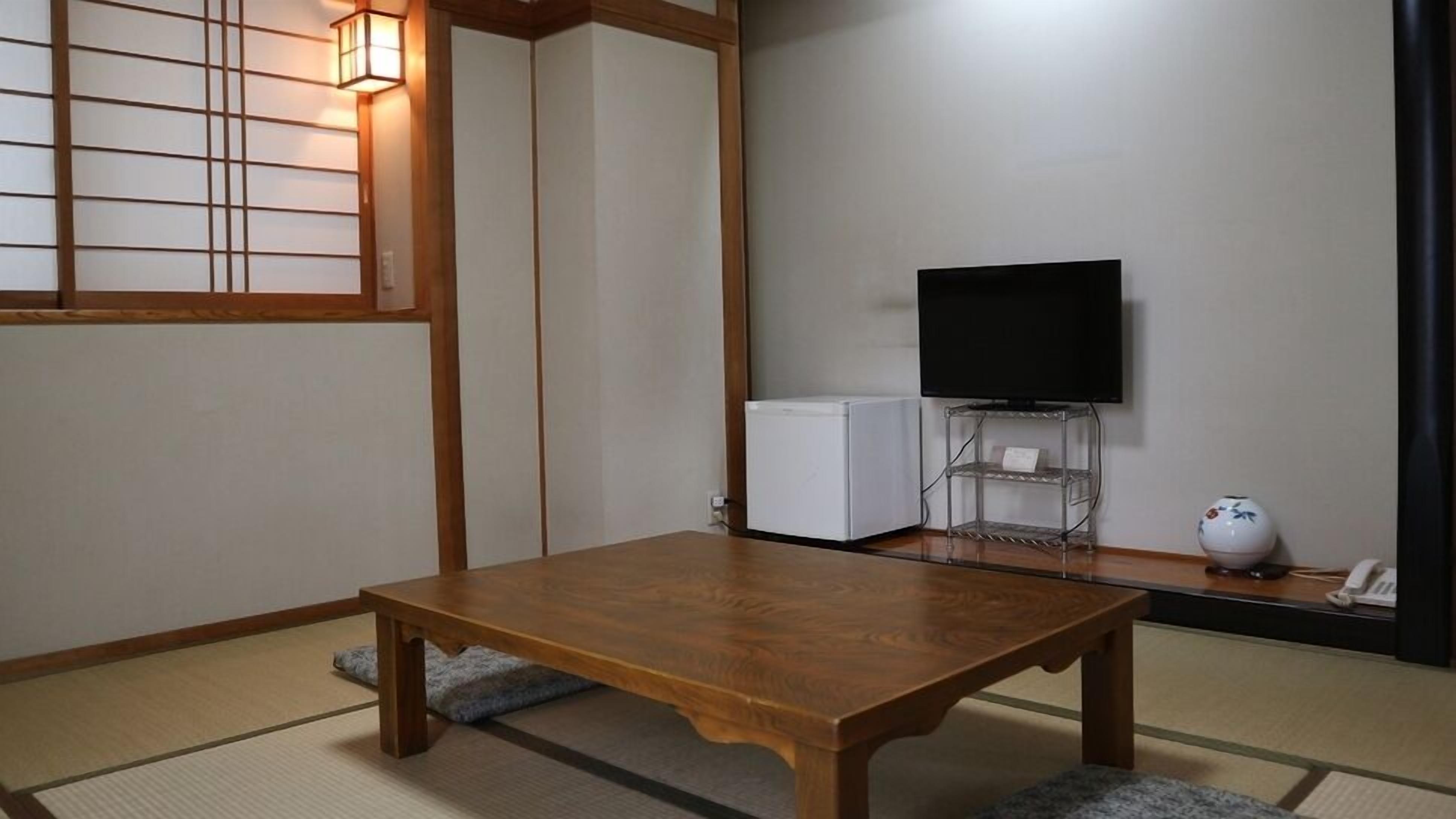 Ein traditioneller japanischer Raum mit Tatami-Böden, einem Holztisch, einem Fernseher auf einem Standfuß und einem kleinen Kühlschrank.