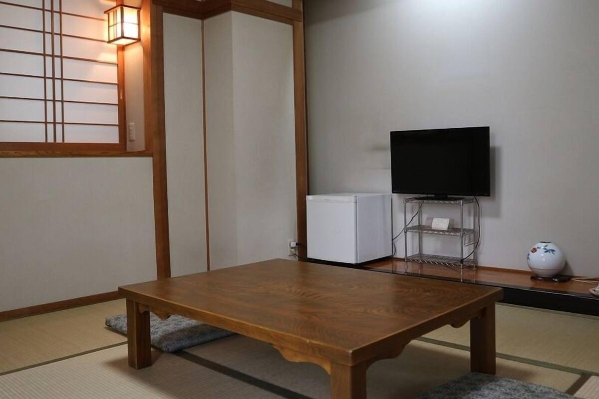 A traditional Japanese room with tatami flooring, a wooden table, a television on a stand, and a small refrigerator.