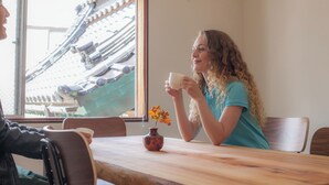 Une personne assise à une table en bois, devant une fenêtre donnant sur des bâtiments d’architecture traditionnelle.