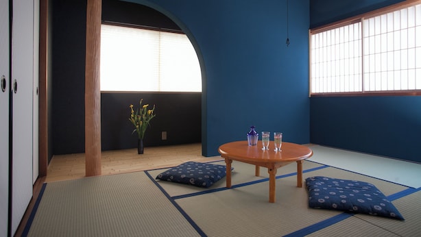 Une pièce avec un sol recouvert de tatamis, une table en bois, des coussins, un vase de fleurs et une grande fenêtre avec des stores.