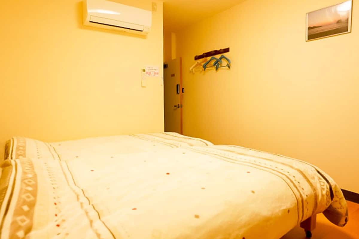 A hotel room with a bed, an air conditioning unit, a door, and a wall-mounted rack with clothes.