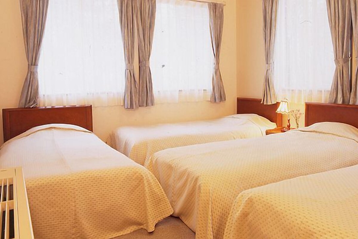 A hotel room with three single beds, a wooden headboard, a nightstand with a lamp, and curtains on the windows.