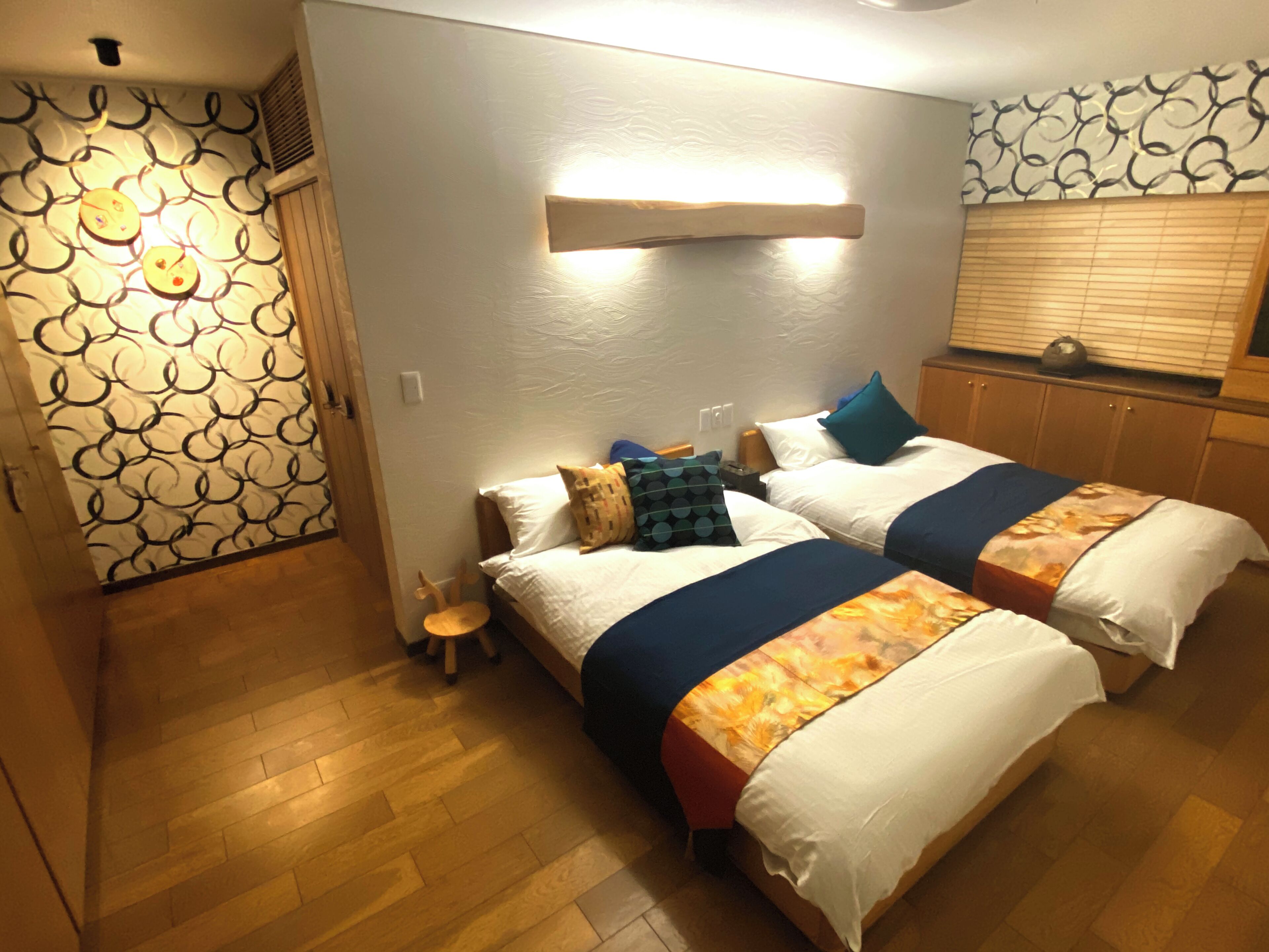 A hotel room with two beds, wooden flooring, a small side table, a wall-mounted light, and a patterned wall.