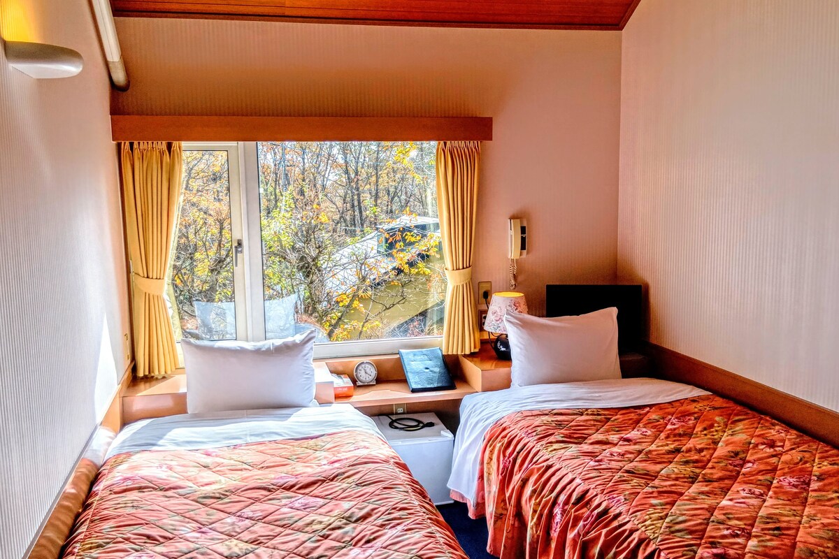 A hotel room with two beds, a window with curtains, a bedside table with a lamp, a clock, and a view of trees outside.