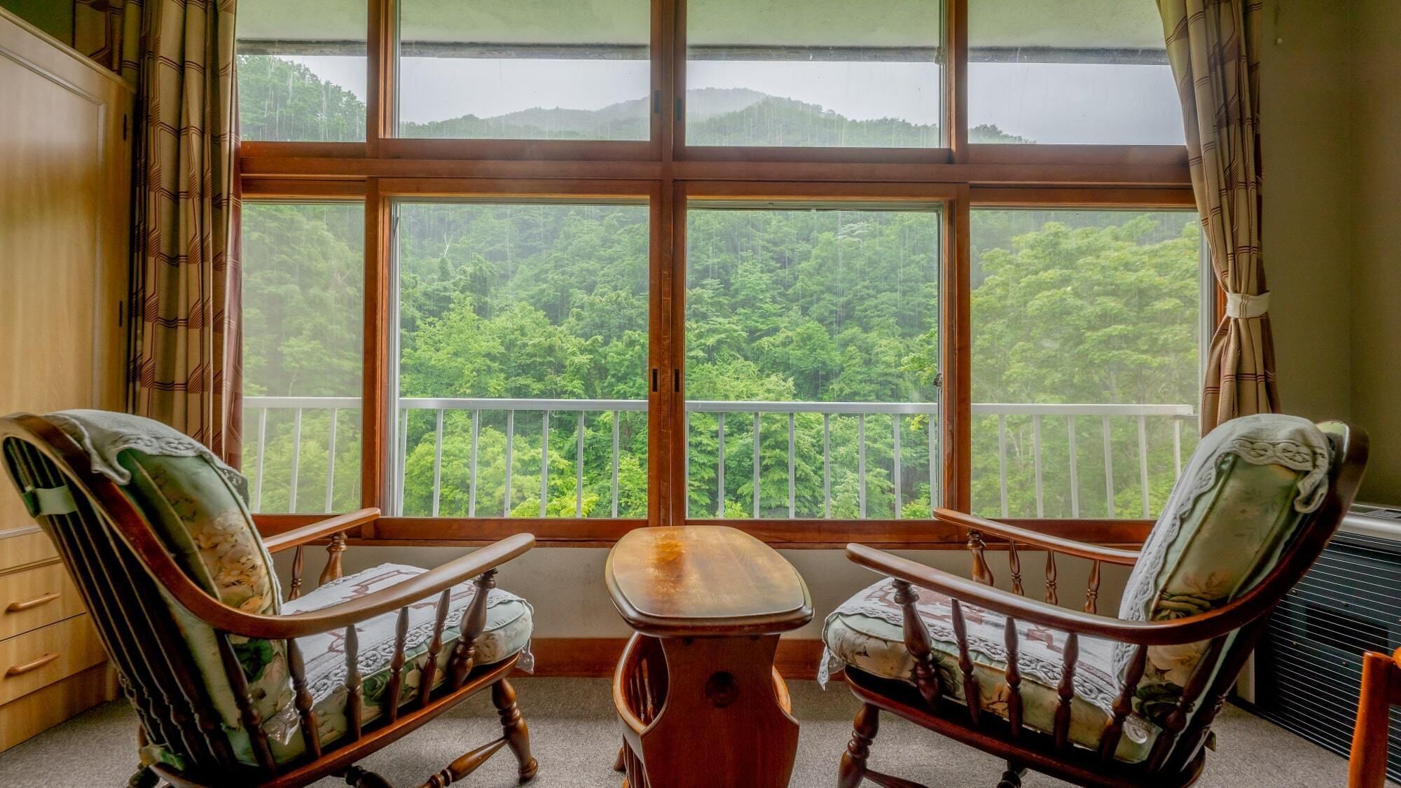 A room with two wooden chairs, a small table, and a large window with a view of a forested area.