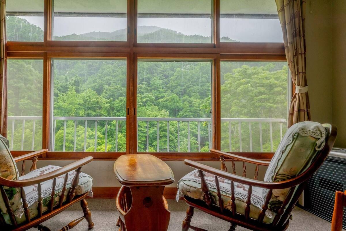 Ein Zimmer mit zwei Holzsesseln, einem kleinen Tisch und einem großen Fenster mit Blick auf ein bewaldetes Gebiet.