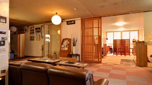 Ein traditioneller japanischer Raum mit Tatami-Boden, einem niedrigen Tisch mit Büchern, einer Schiebetür und Blick in einen weiteren Raum mit Schreibtisch und Stühlen.