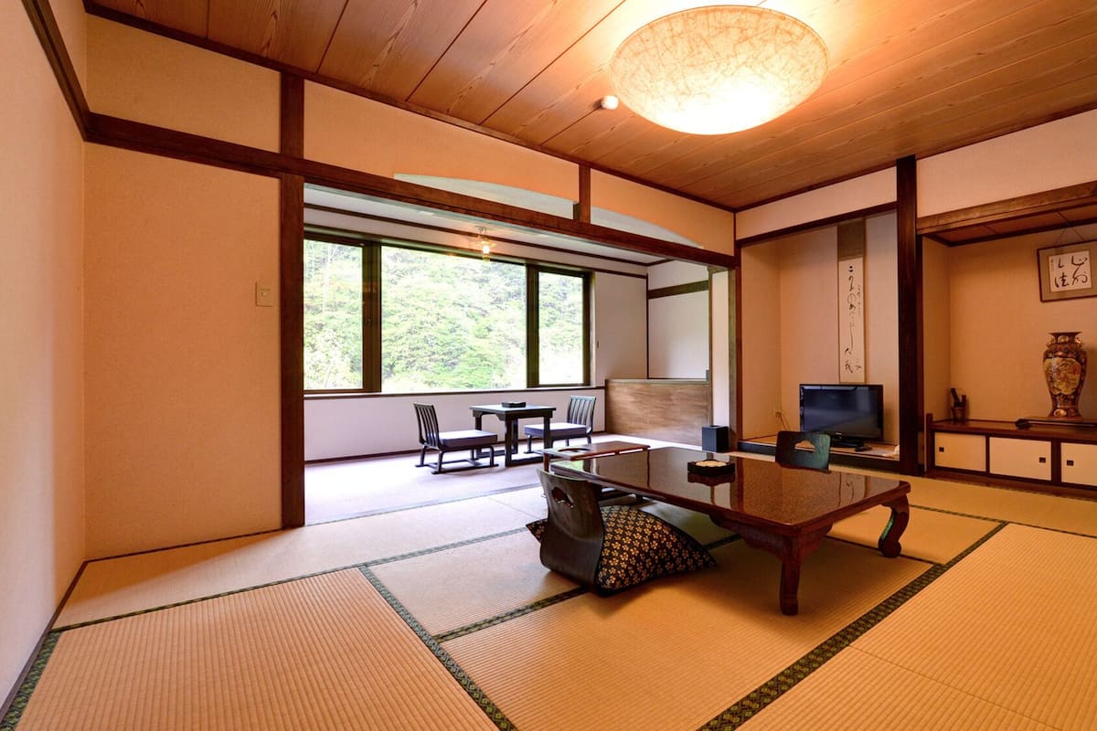 Ein traditioneller japanischer Raum mit Tatami-Bodenbelag, einem niedrigen Tisch, einem Fernseher und Blick auf Grünflächen.